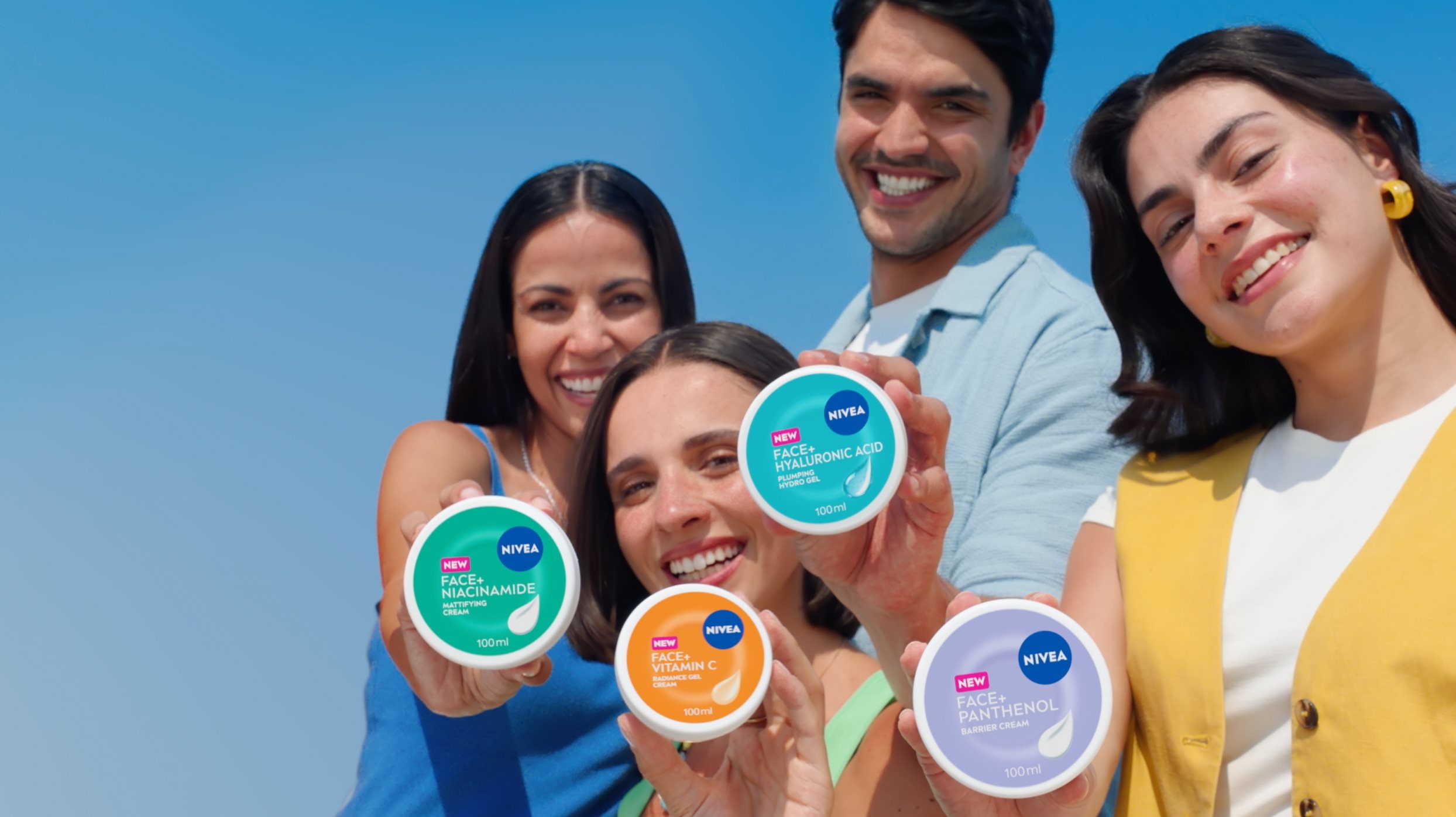 People smiling and showing NIVEA FACE+ cream jars in turquoise, orange, blue, and violet, each featuring the NIVEA FACE+ logo and a distinctive color-coded design.